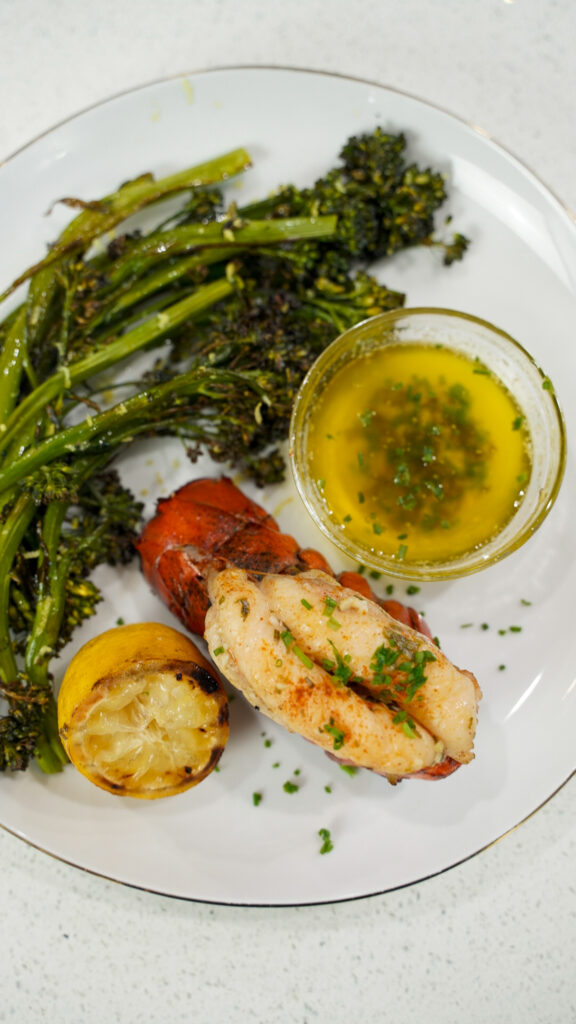 Grilled lobster tail on a plate with garlic herb butter and broccolini.